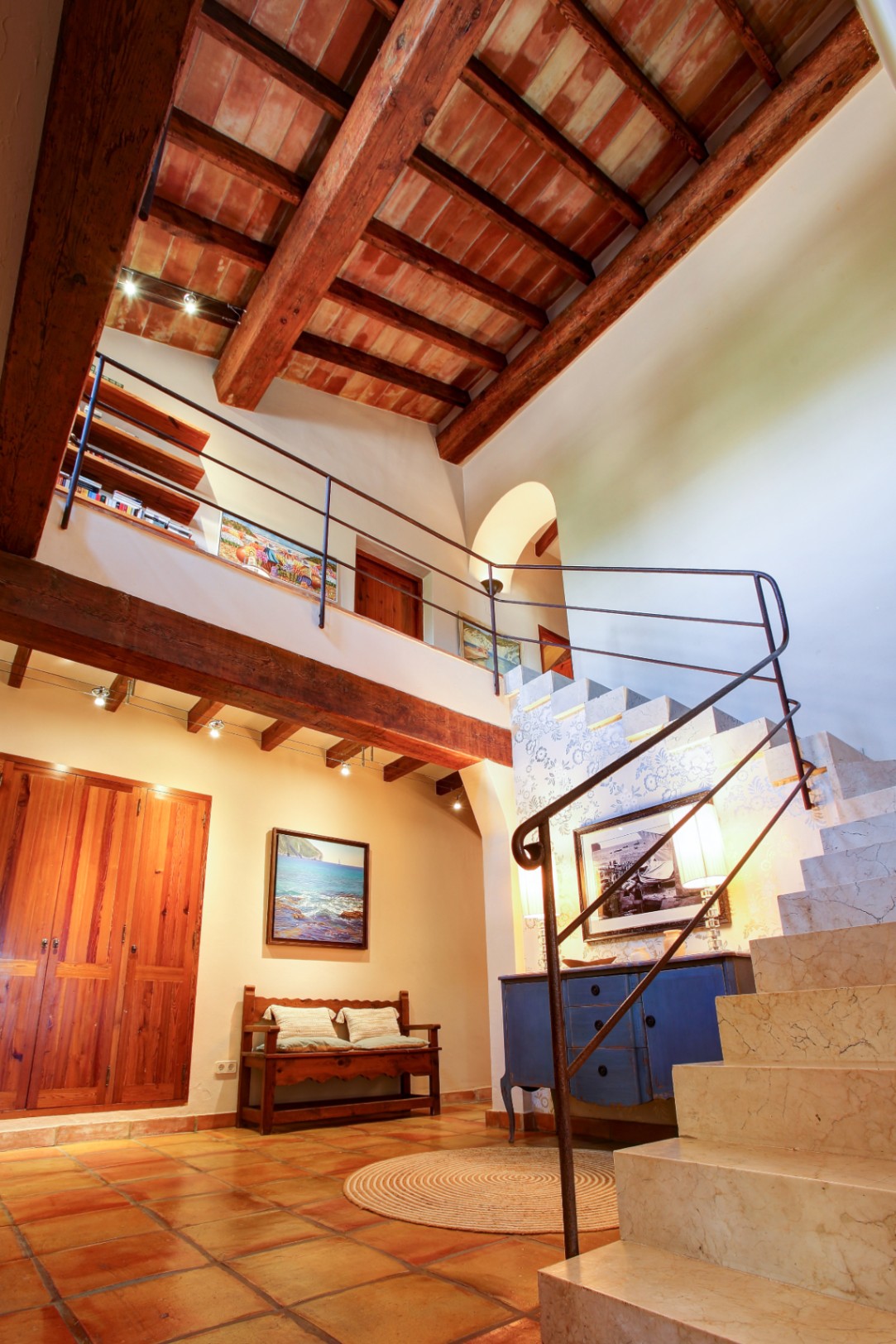 Entrance hall featuring double-height ceilings with exposed wooden beams and terracotta tiles. A solid marble staircase with wrought iron railings leads to the upper mezzanine.