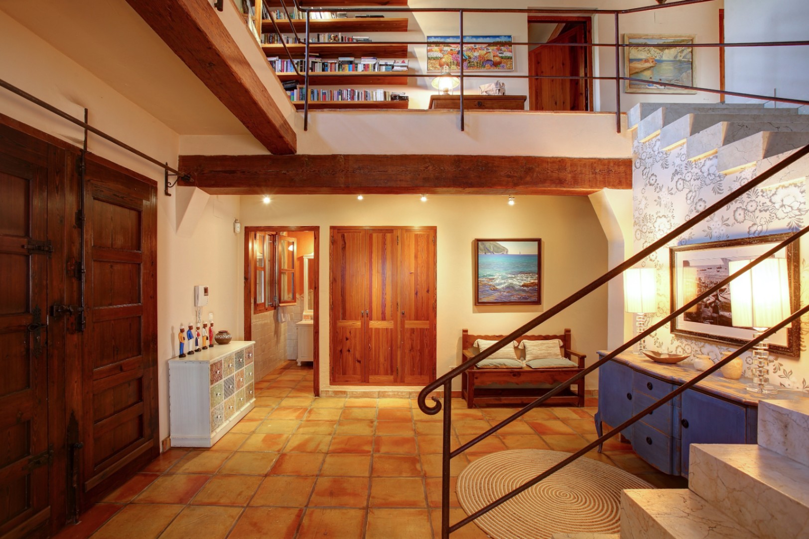 Entrance hall featuring terracotta tile flooring, exposed wooden beams, and a marble staircase leading to an upper mezzanine library with integrated shelving.