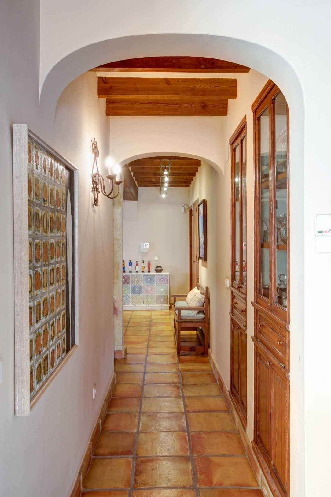 Hallway featuring arched entryways, exposed wooden ceiling beams, and traditional terracotta flooring. Includes built-in wooden display cabinets and rustic wall lighting.