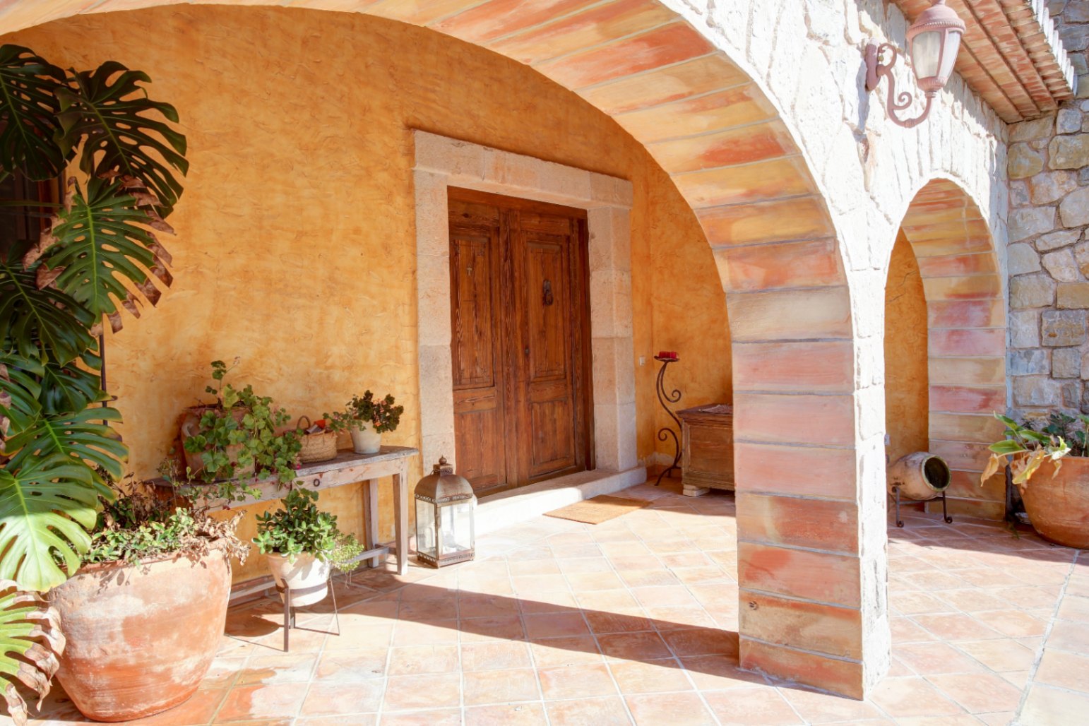 Main entrance featuring a rustic stone-arched porch, terracotta tile flooring, a solid wood double door, and lush Mediterranean potted plants.