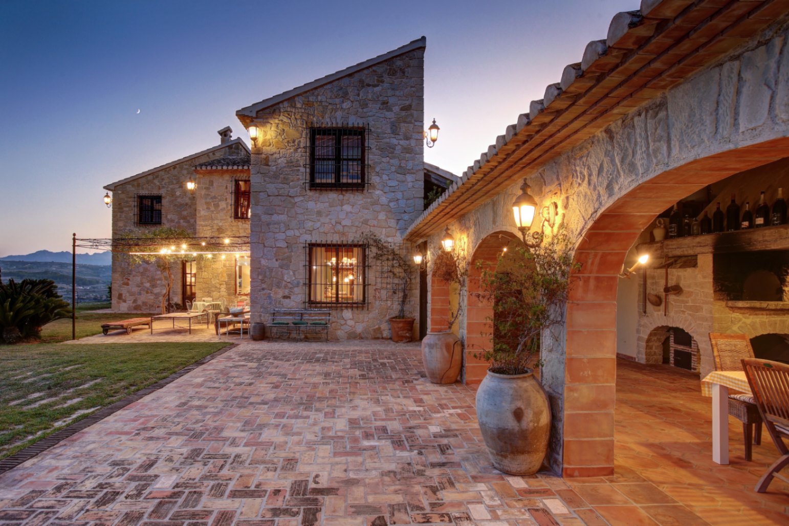 Natural stone facade and terracotta courtyard at dusk. Featuring exposed brick arches, an outdoor kitchen area, and warm lantern lighting against a twilight sky.