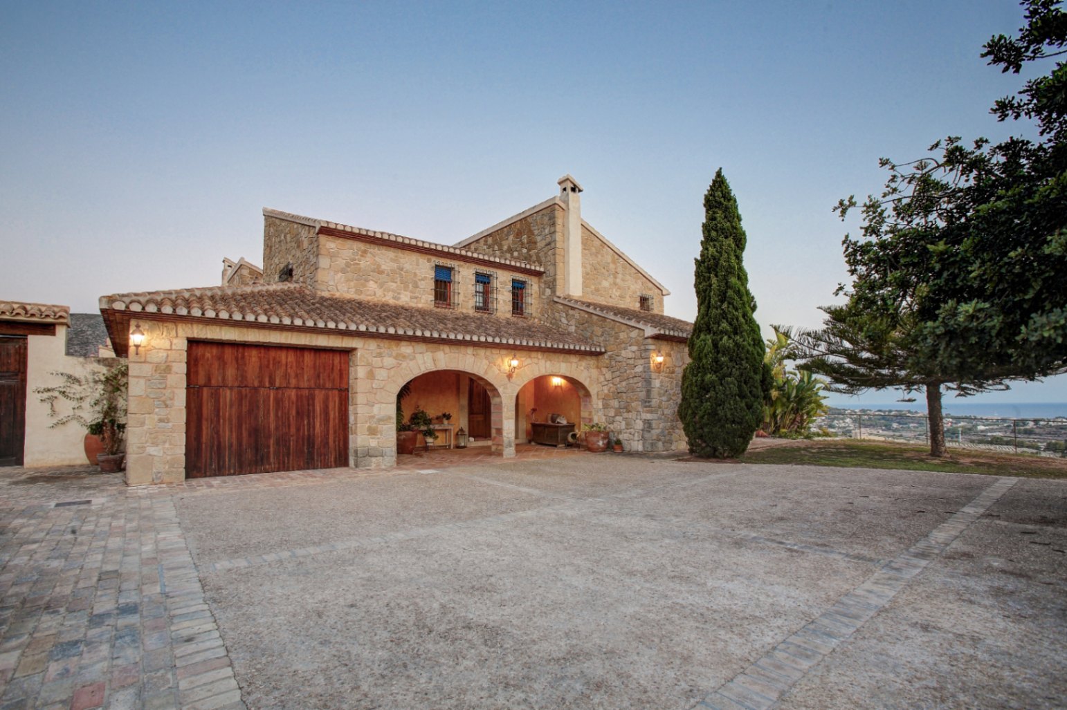 Natural stone facade featuring rustic arches, a solid wood garage door, and a spacious paved courtyard with distant Mediterranean Sea views.