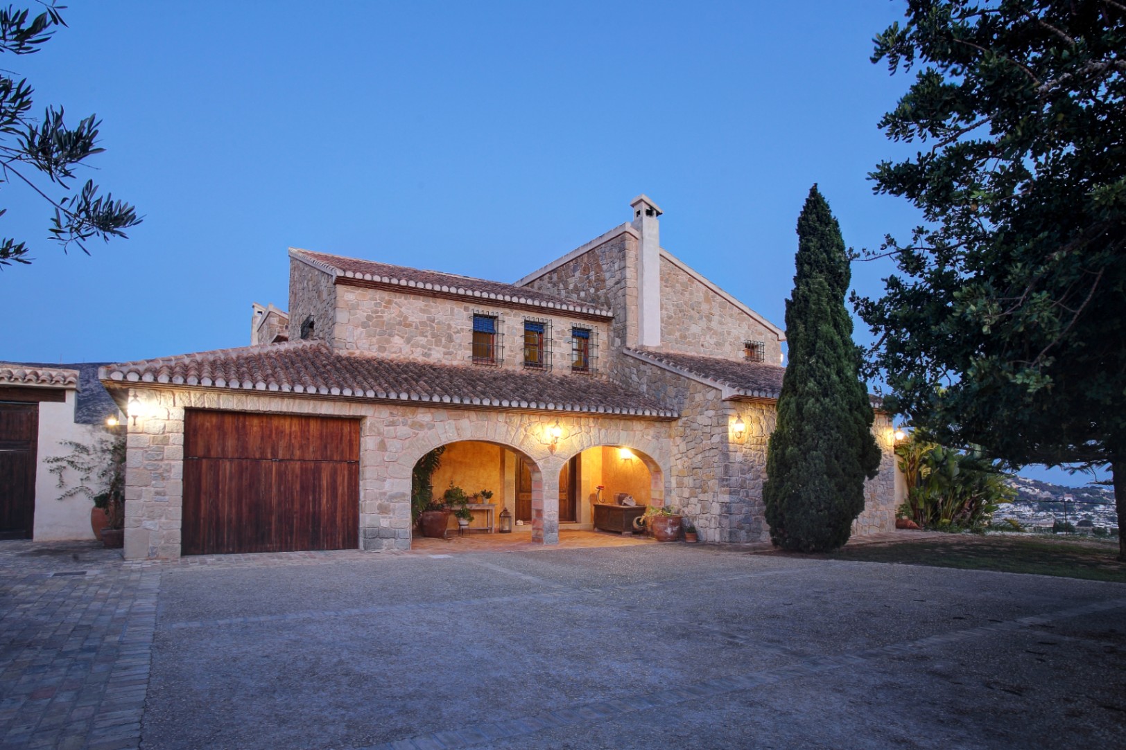 Natural stone facade featuring traditional arches and a wooden garage door, captured at dusk with warm exterior lighting and a paved driveway.