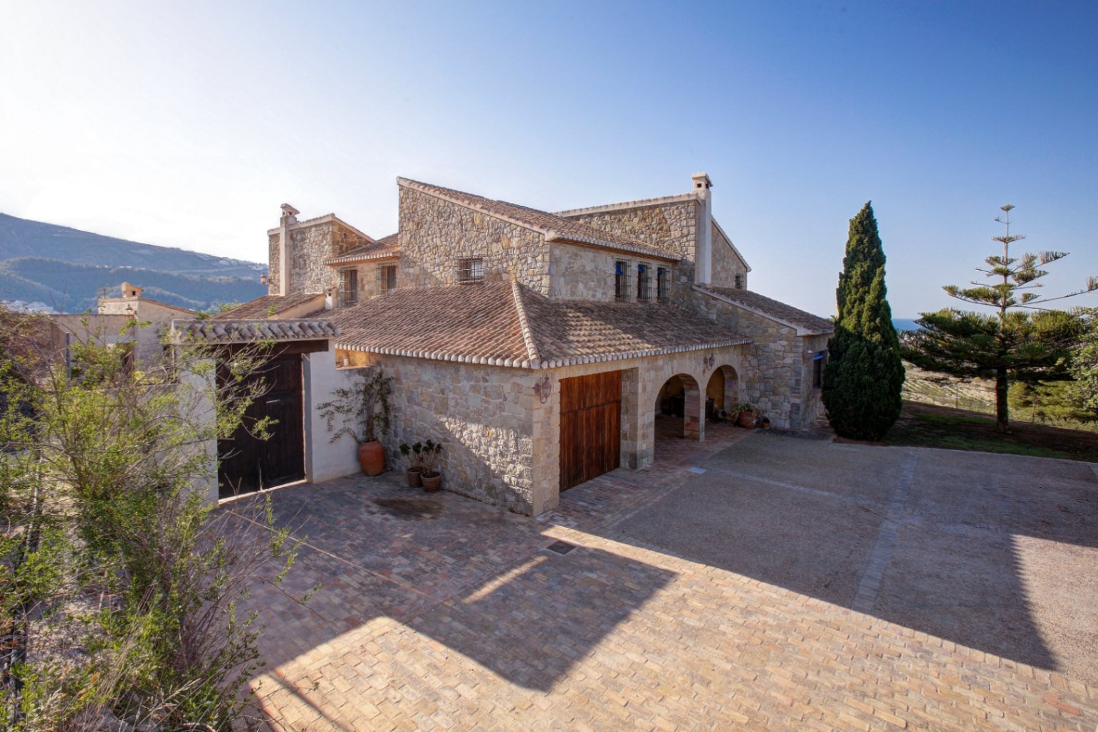 Natural stone facade featuring traditional arches, clay tile roofing, and a spacious terracotta paved courtyard under the Mediterranean sun.