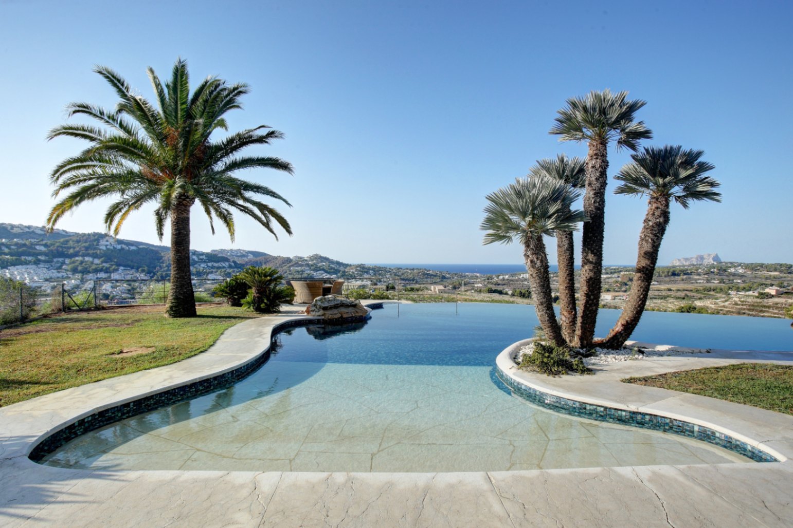 Organic-shaped infinity pool framed by mature palm trees, offering panoramic views of the Mediterranean Sea and the surrounding coastal landscape under a clear sky.