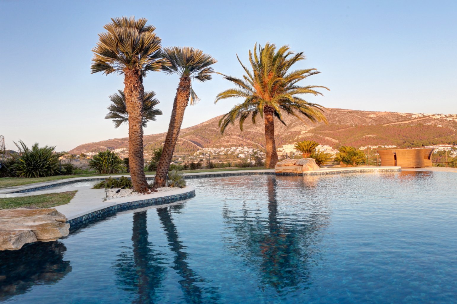 Organic-shaped pool featuring dark blue mosaic tiling and integrated palm trees, overlooking a scenic valley and mountain range during the golden hour.