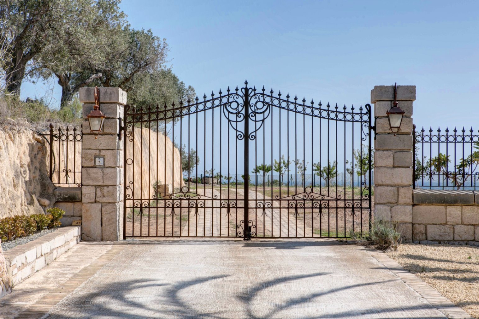 Ornate wrought-iron entrance gate set between natural stone pillars with traditional lanterns, leading to a paved driveway under a clear sky.