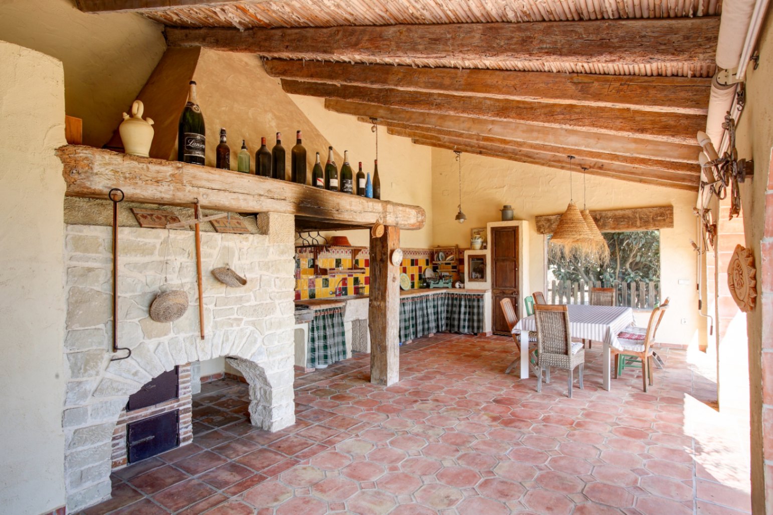 Outdoor summer kitchen featuring a large stone fireplace, reclaimed wooden beams, terracotta flooring, and a rustic dining area with woven lamps.
