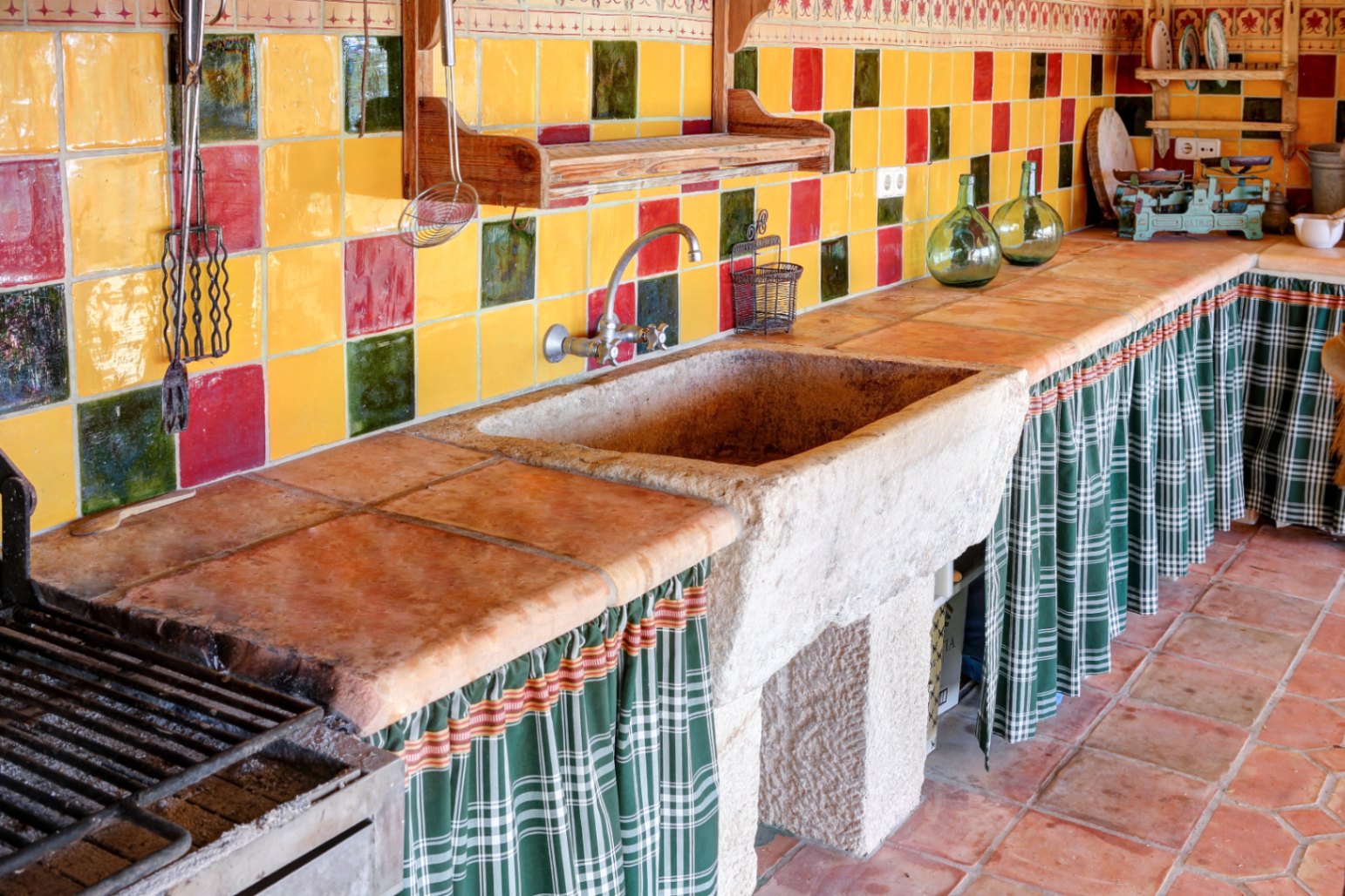 Outdoor summer kitchen featuring a solid stone sink, terracotta tile countertops, and a vibrant ceramic backsplash. Includes a built-in grill and traditional under-counter curtains.