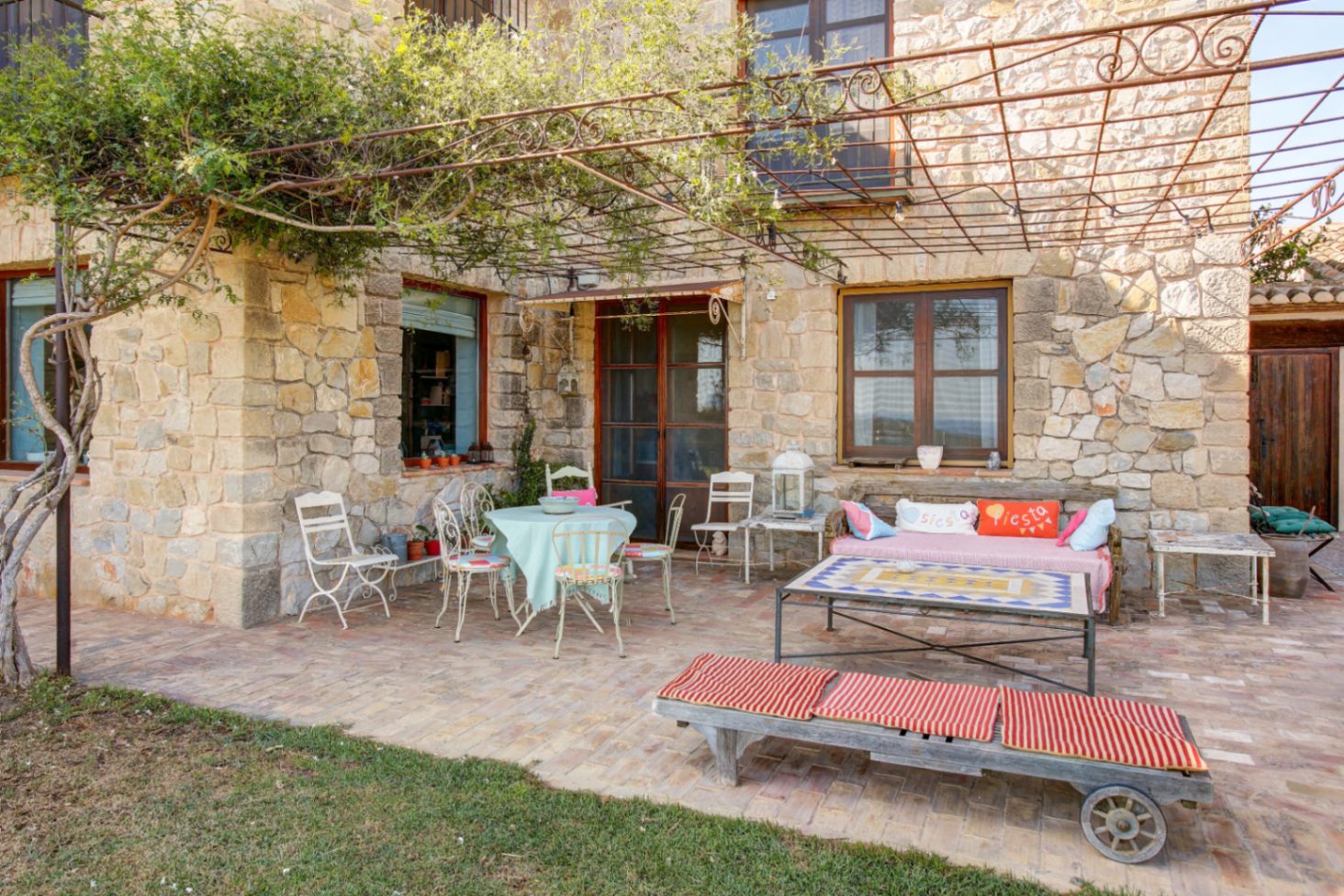 Outdoor terrace featuring a natural stone facade, terracotta brick flooring, and a wrought iron pergola. The space includes rustic seating areas and a mosaic-topped coffee table.