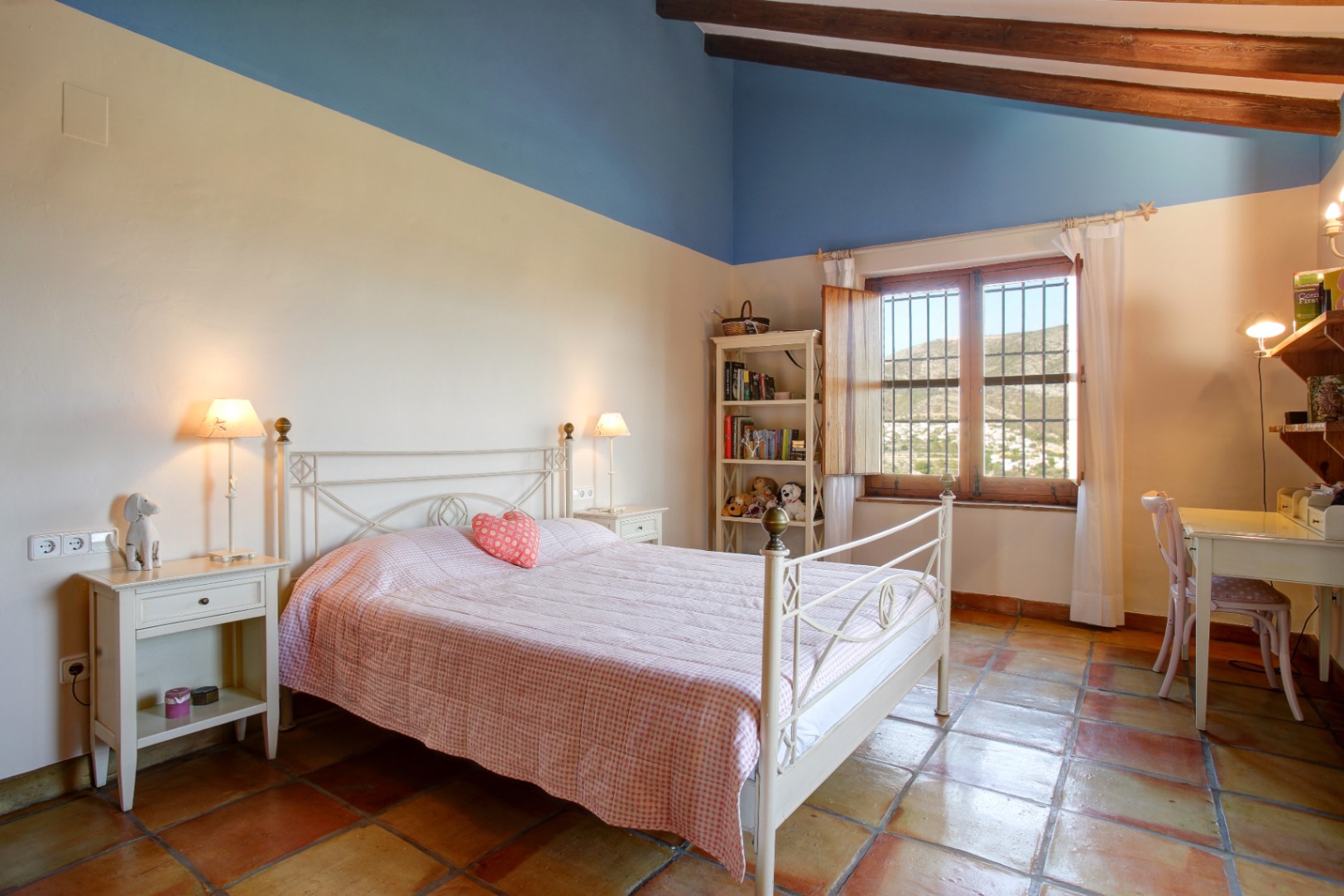 Rustic bedroom featuring exposed wooden beams, terracotta tile flooring, and a white wrought iron bed. The space includes a study desk and views of the surrounding hills.