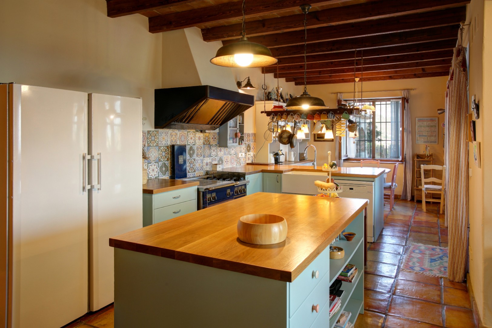 Rustic kitchen featuring a wooden island, exposed ceiling beams, and terracotta flooring. Includes a professional blue range stove, decorative tile backsplash, and ample natural light.