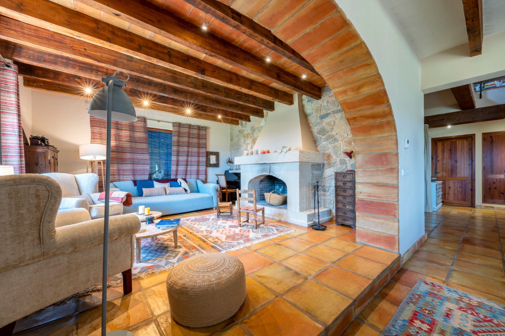 Rustic living area featuring a stone fireplace, exposed wooden beams with terracotta ceiling tiles, traditional clay floors, and a prominent brick archway.