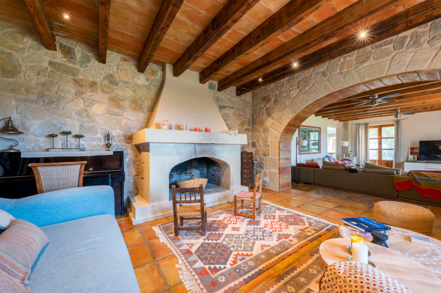 Rustic living area featuring natural stone walls, a traditional fireplace, and exposed timber beams. A prominent stone archway connects the lounge spaces over terracotta tiling.