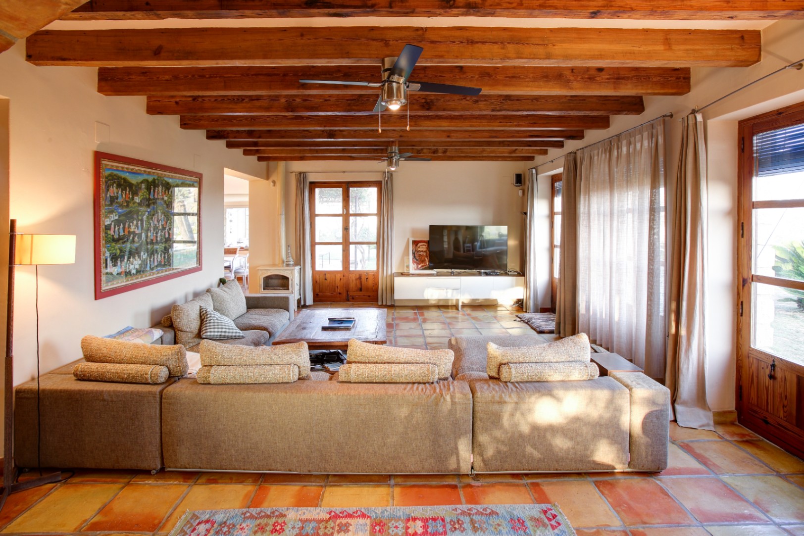 Rustic living room featuring exposed wooden beams, terracotta tile flooring, a white wood-burning stove, and large wooden-framed doors leading outside.