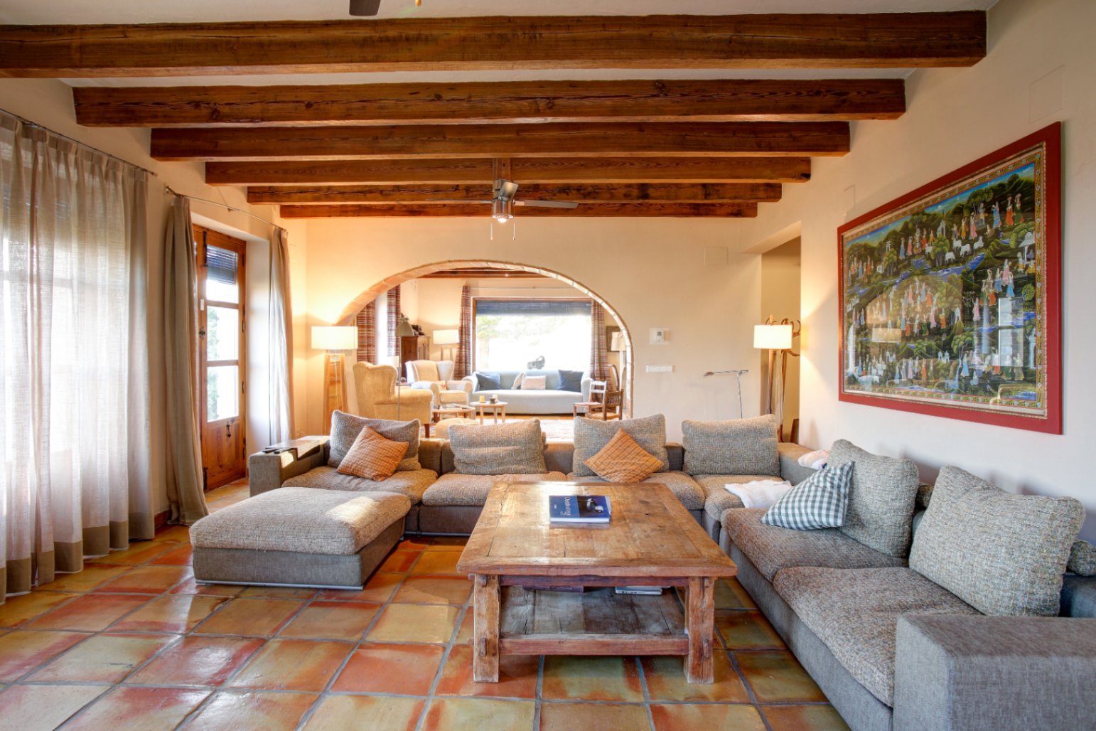 Rustic living room featuring exposed wooden beams, terracotta tile flooring, and a large modular sofa. A wide archway connects to a secondary sunlit seating area.