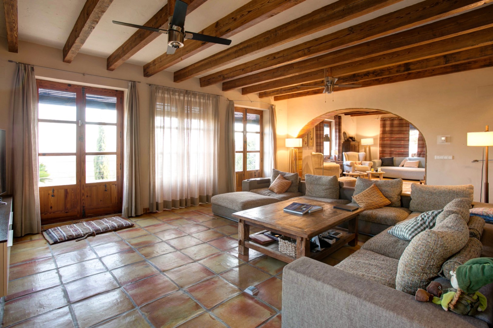 Rustic living room featuring terracotta tile flooring, exposed wooden ceiling beams, and a large archway. Wooden French doors provide abundant natural light and outdoor access.