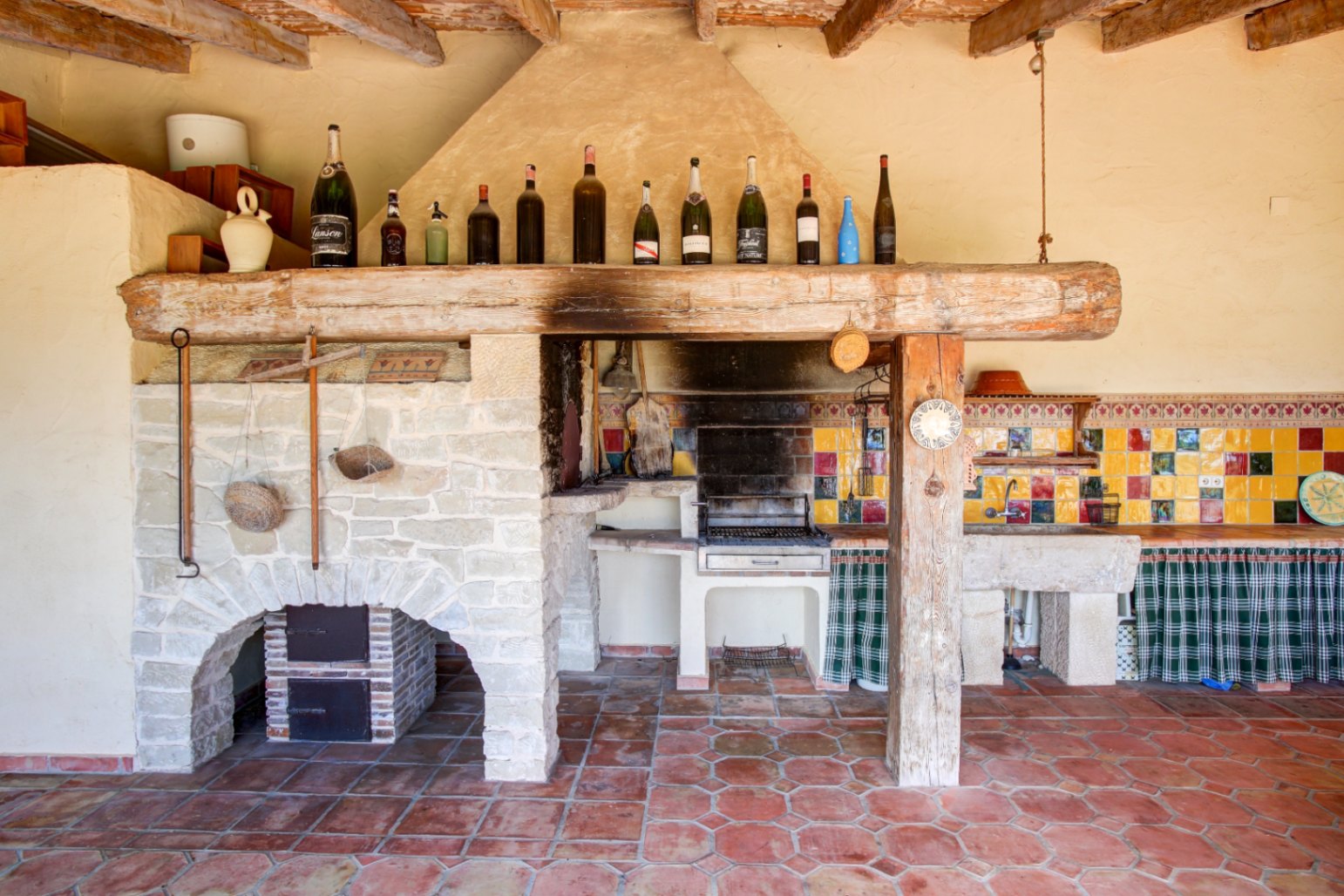 Rustic summer kitchen featuring a built-in barbecue, massive reclaimed wood lintel, natural stone sink, and vibrant ceramic tiling over hexagonal terracotta flooring.