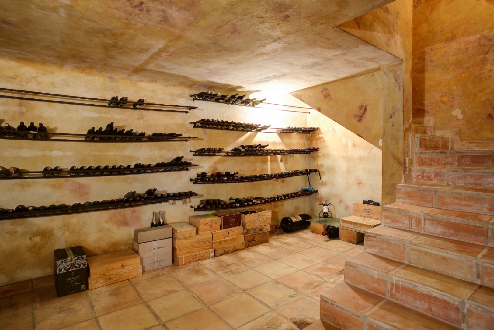 Rustic wine cellar featuring terracotta tile flooring, textured plaster walls, and wall-mounted metal bottle racks beneath a staircase.