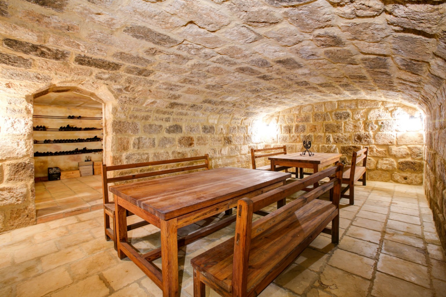 Rustic wine cellar with vaulted stone ceilings, terracotta tile flooring, and solid wood dining benches, featuring an adjacent wine storage room.