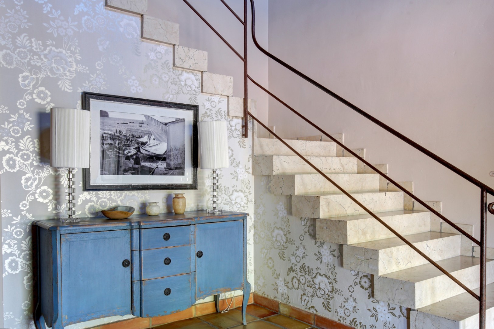Solid marble staircase with a metal handrail, featuring a blue distressed sideboard, floral wallpaper, and traditional terracotta floor tiles.