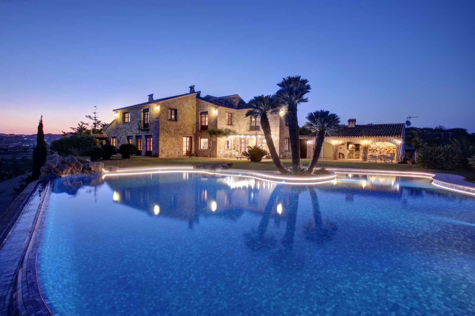 Twilight view of the natural stone facade and organic-shaped pool with integrated LED lighting, featuring the outdoor kitchen and traditional barbecue area.