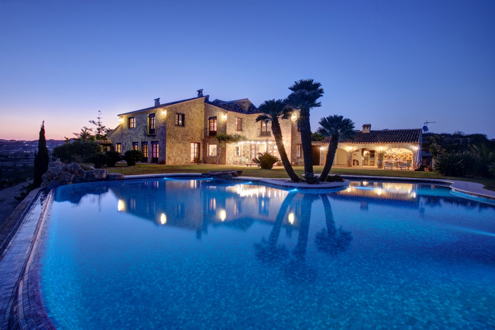 Twilight view of the stone facade and illuminated pool, featuring the outdoor kitchen and palm trees integrated into the Mediterranean landscape.