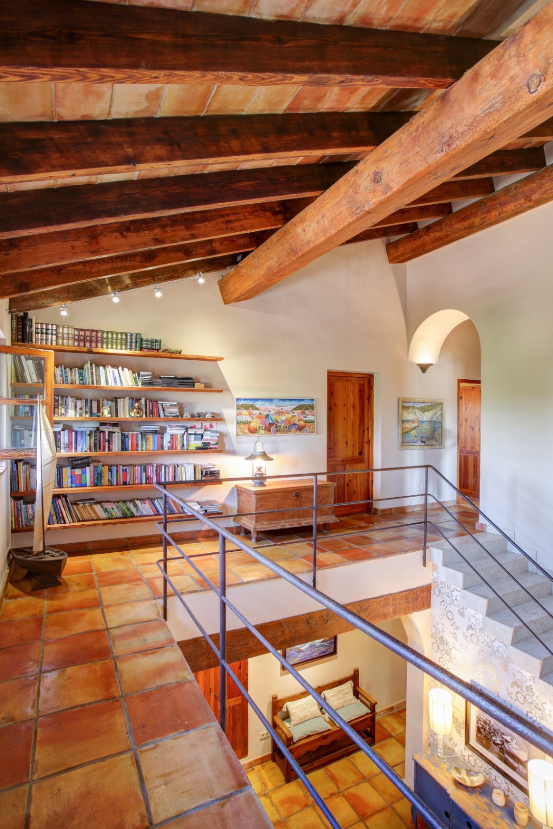 Upper floor landing featuring exposed wooden beams, terracotta tile flooring, a built-in library, and a wrought iron railing overlooking the lower level.