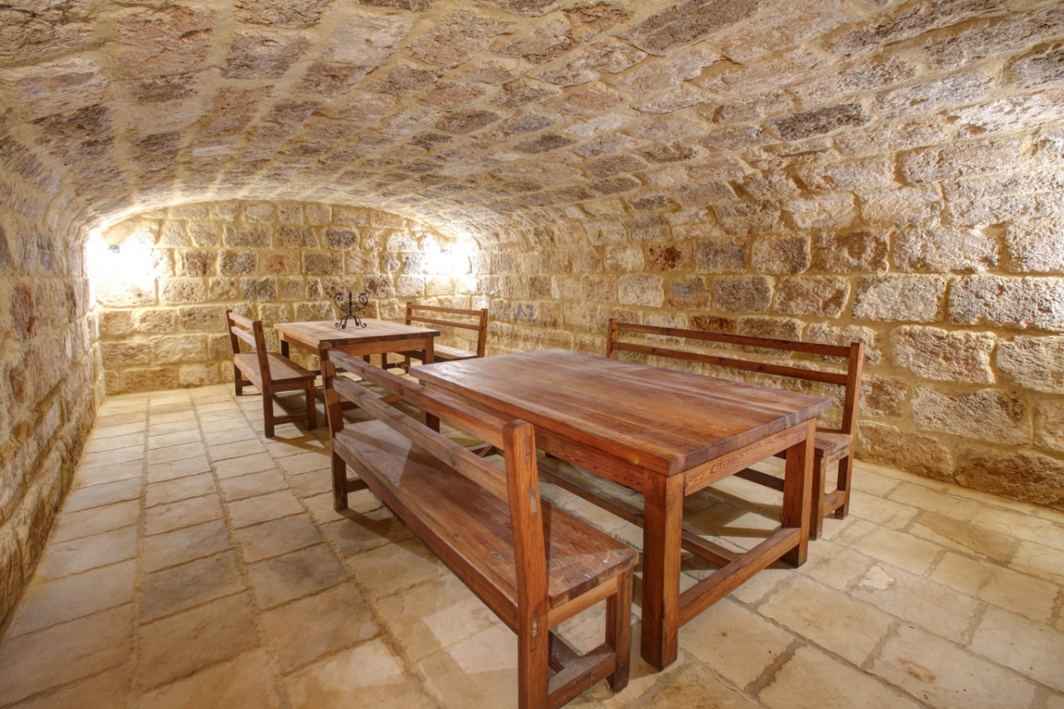 Vaulted stone cellar featuring traditional masonry, solid wooden dining sets, and rustic stone flooring, illuminated by warm wall-mounted lighting.