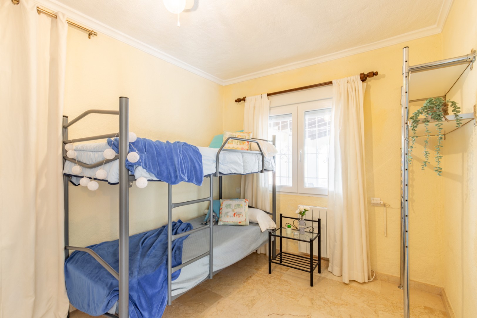 Bedroom featuring a grey metal bunk bed, cream walls, and polished stone flooring. The space includes a bright window and an open metal shelving unit.