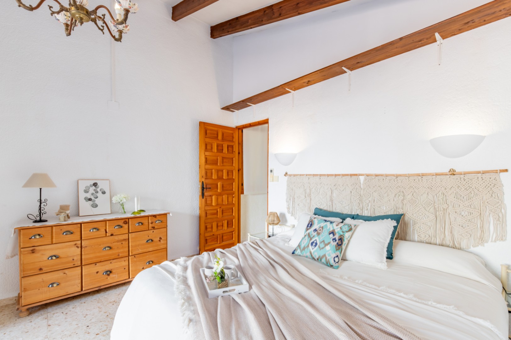 Bedroom featuring exposed wooden ceiling beams, a pine dresser, and a macramé headboard. The space includes original terrazzo flooring and a traditional carved wooden door.