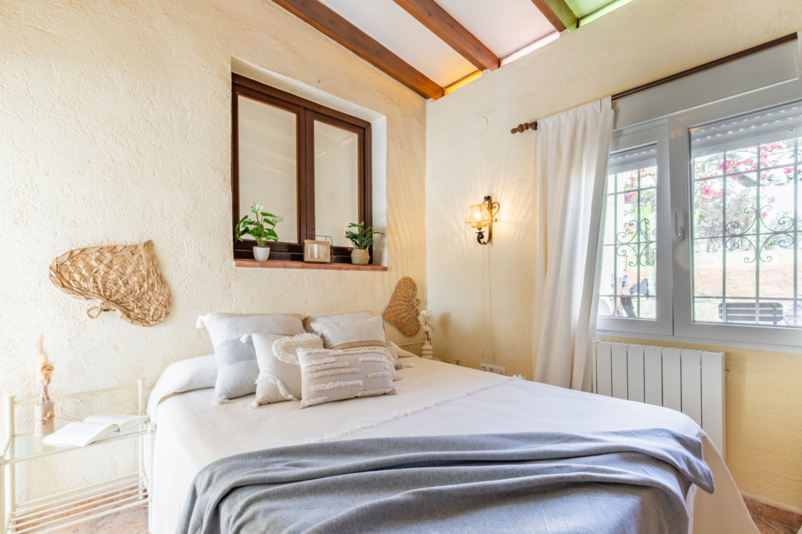 Bedroom featuring exposed wooden ceiling beams, rustic textured walls, and dual windows. The space includes a wall-mounted lamp and traditional Mediterranean decor.