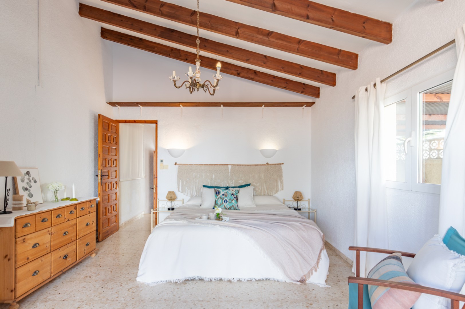 Bedroom featuring high vaulted ceilings with exposed wooden beams, terrazzo flooring, and Mediterranean decor including a macramé headboard and natural wood furniture.