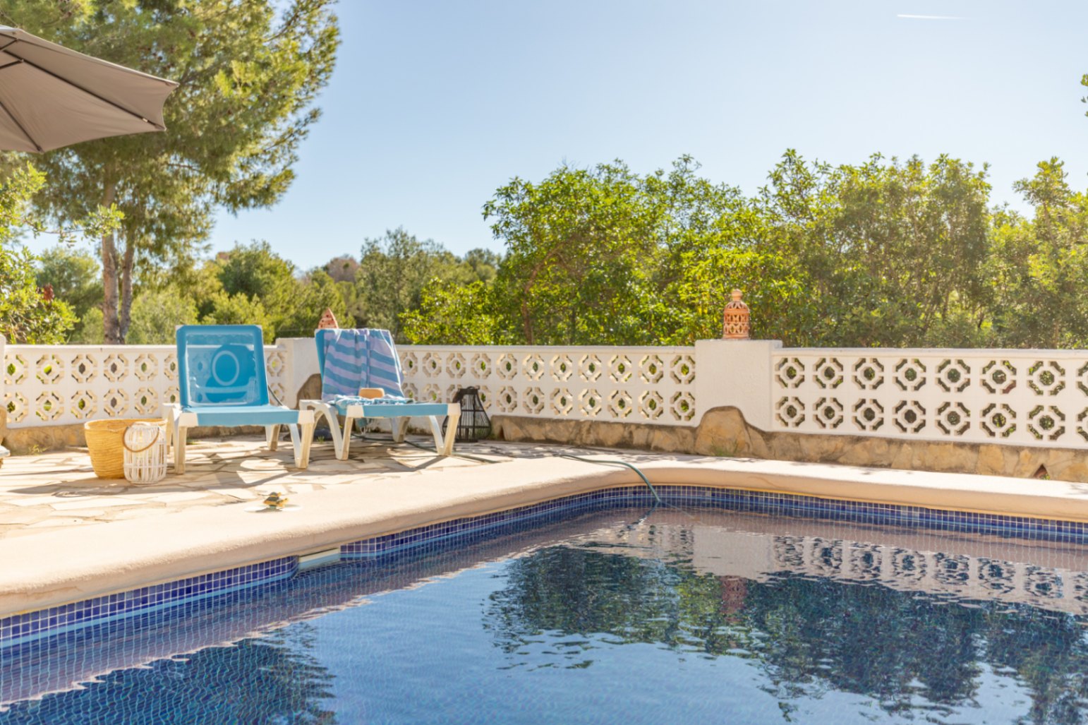 Blue mosaic swimming pool featuring a natural stone sun deck, bordered by a traditional white lattice wall and lush green Mediterranean foliage.
