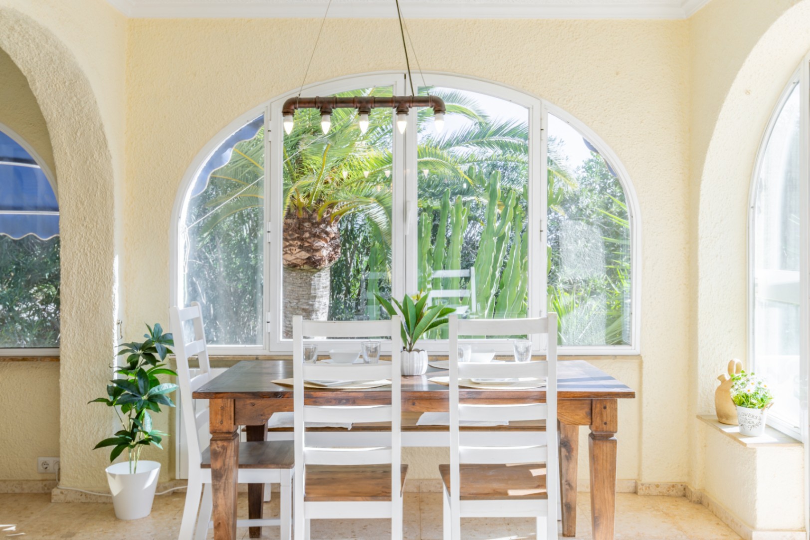 Bright dining area in a glazed naya featuring a wooden table, white chairs, and industrial lighting with views of the tropical garden.