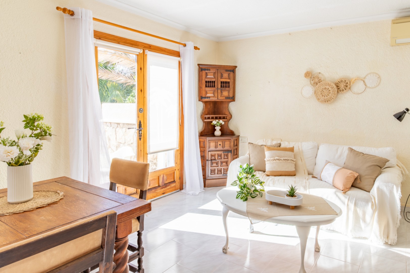 Bright living and dining area featuring rustic wooden furniture, a white sofa, and a large glass door providing direct access to the terrace and natural light.