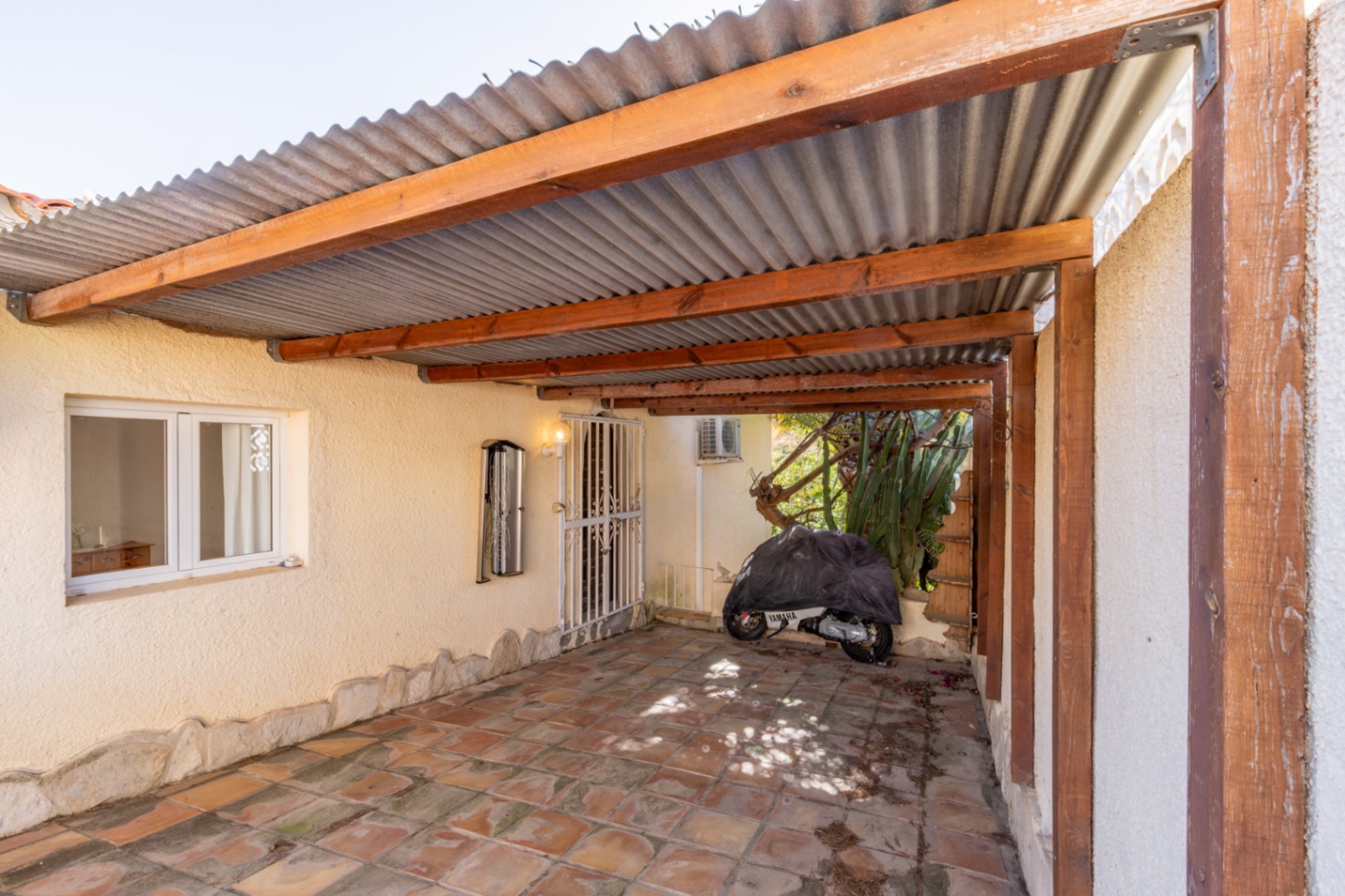 Covered parking area featuring a wooden beam structure, corrugated roofing, and rustic tiled flooring, providing shaded space for vehicles and direct house access.