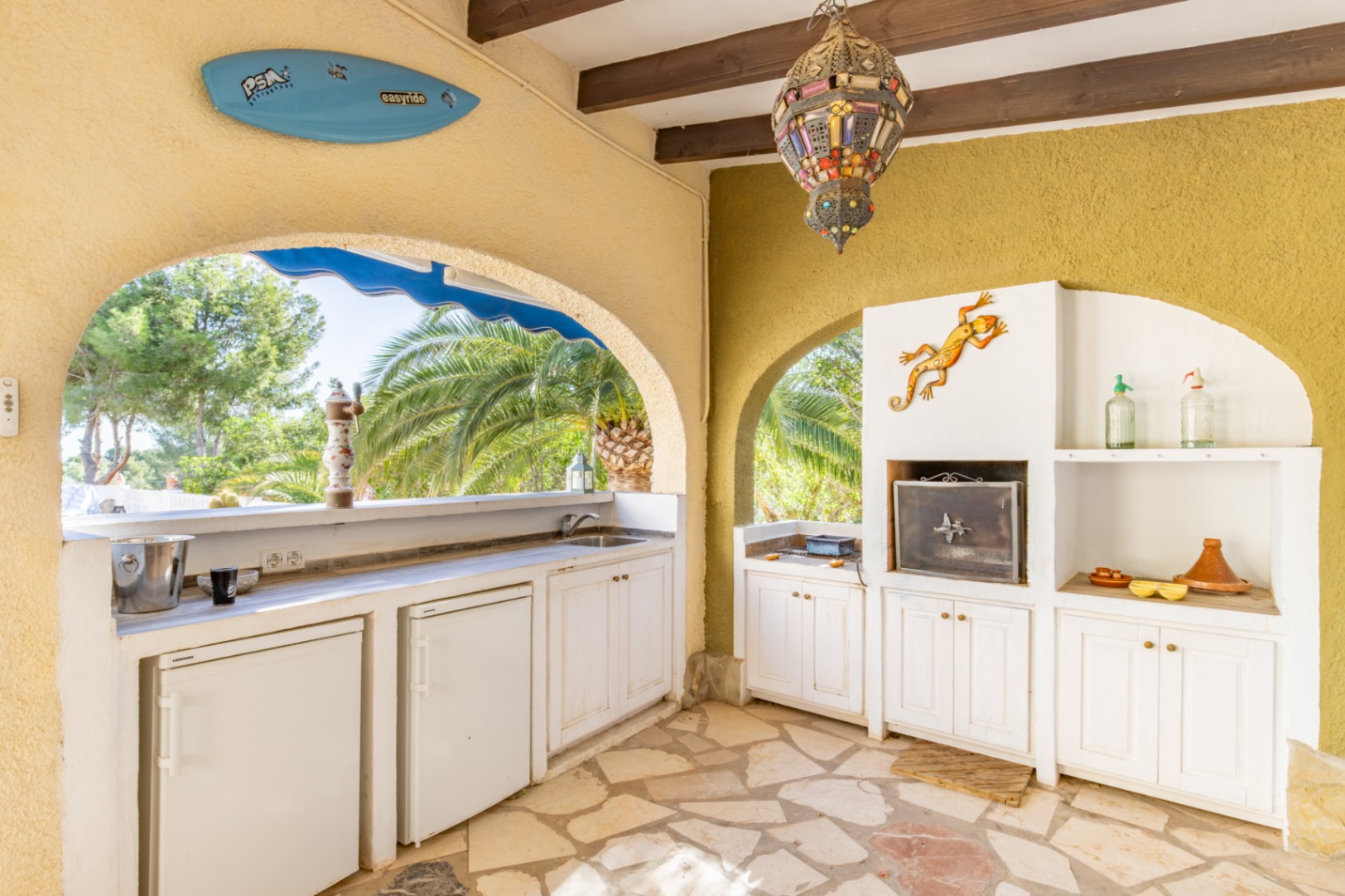 Covered summer kitchen featuring built-in BBQ, multiple under-counter refrigerators, and wooden ceiling beams. The space includes natural stone flooring and views of palm trees.