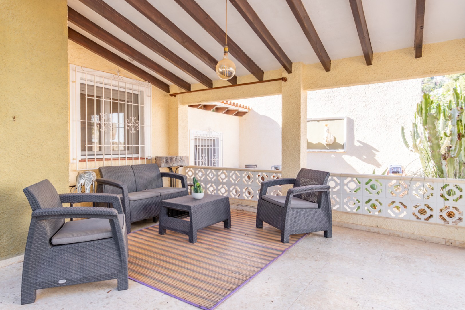Covered terrace featuring exposed wooden ceiling beams, grey wicker-style lounge furniture, and a decorative geometric balustrade overlooking the garden.