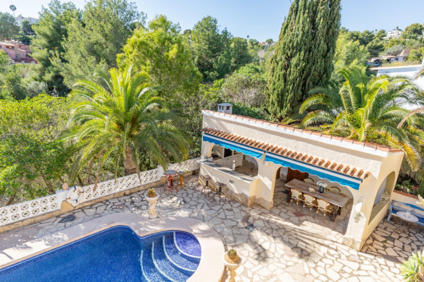 Elevated view of the kidney-shaped pool and stone terrace, featuring a covered summer kitchen and dining area set against a lush, protected green backdrop.