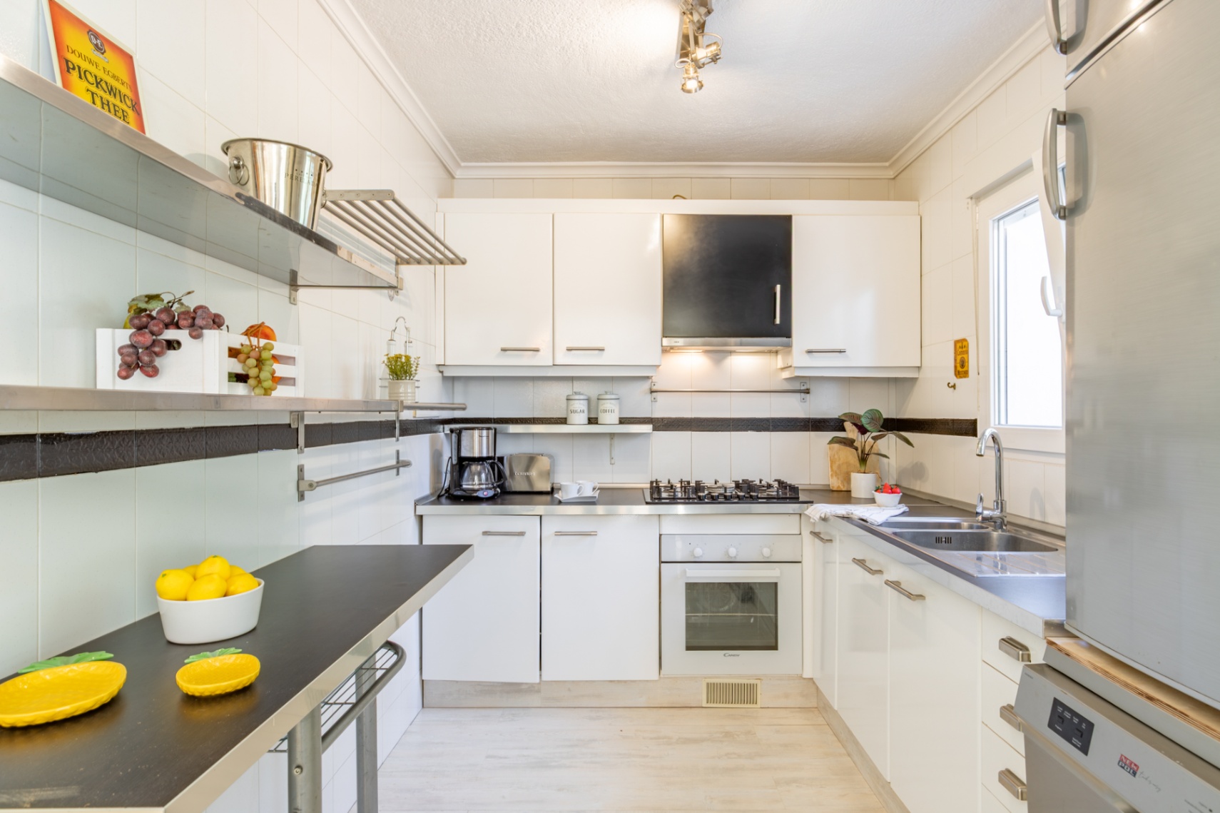 Equipped kitchen featuring white cabinetry, dark countertops, stainless steel shelving, and natural light. Includes a gas hob, oven, and double stainless steel sink.