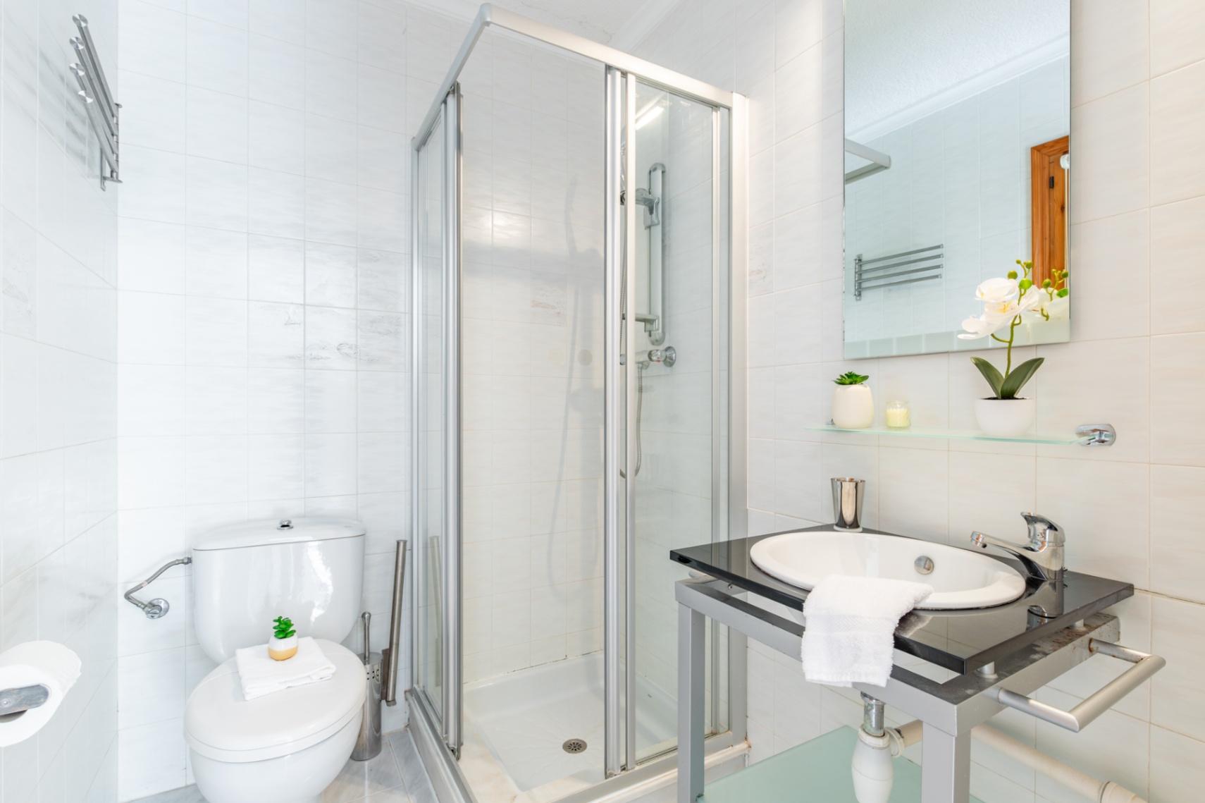Functional bathroom featuring white tiled walls, a corner shower with glass enclosure, and a vanity unit with a dark stone countertop and integrated sink.