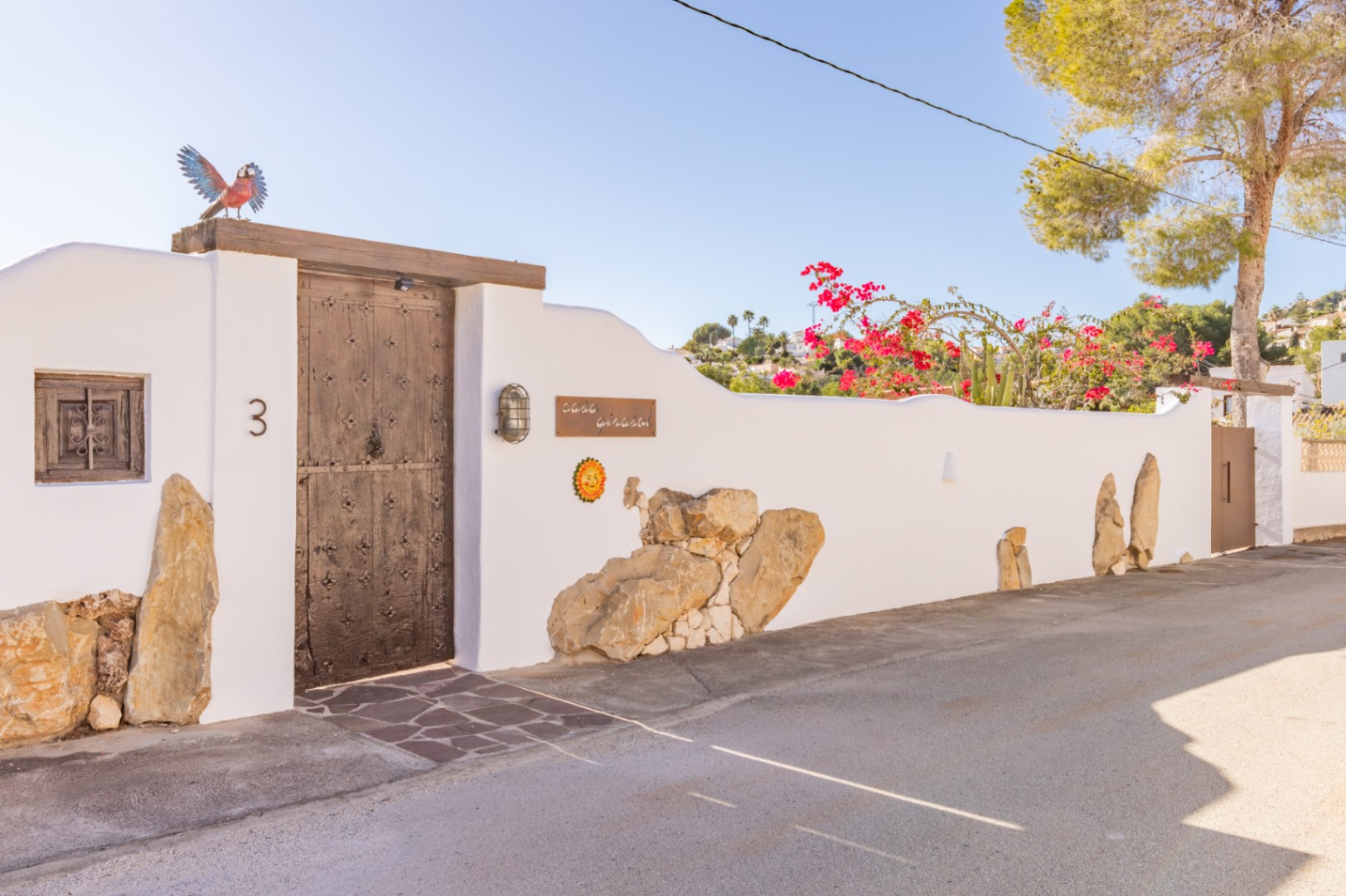 Main entrance featuring a rustic wooden door with iron studs, set in a white perimeter wall with natural stone accents and vibrant bougainvillea.