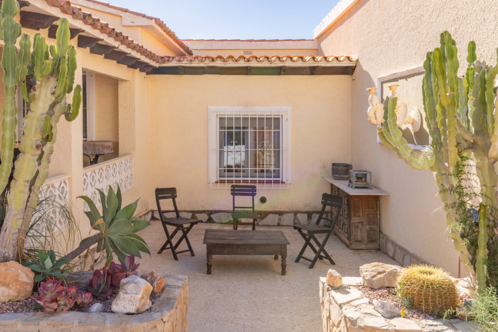 Mediterranean-style outdoor patio featuring stone flooring, rustic wooden furniture, and integrated stone planters with mature cacti and succulents.
