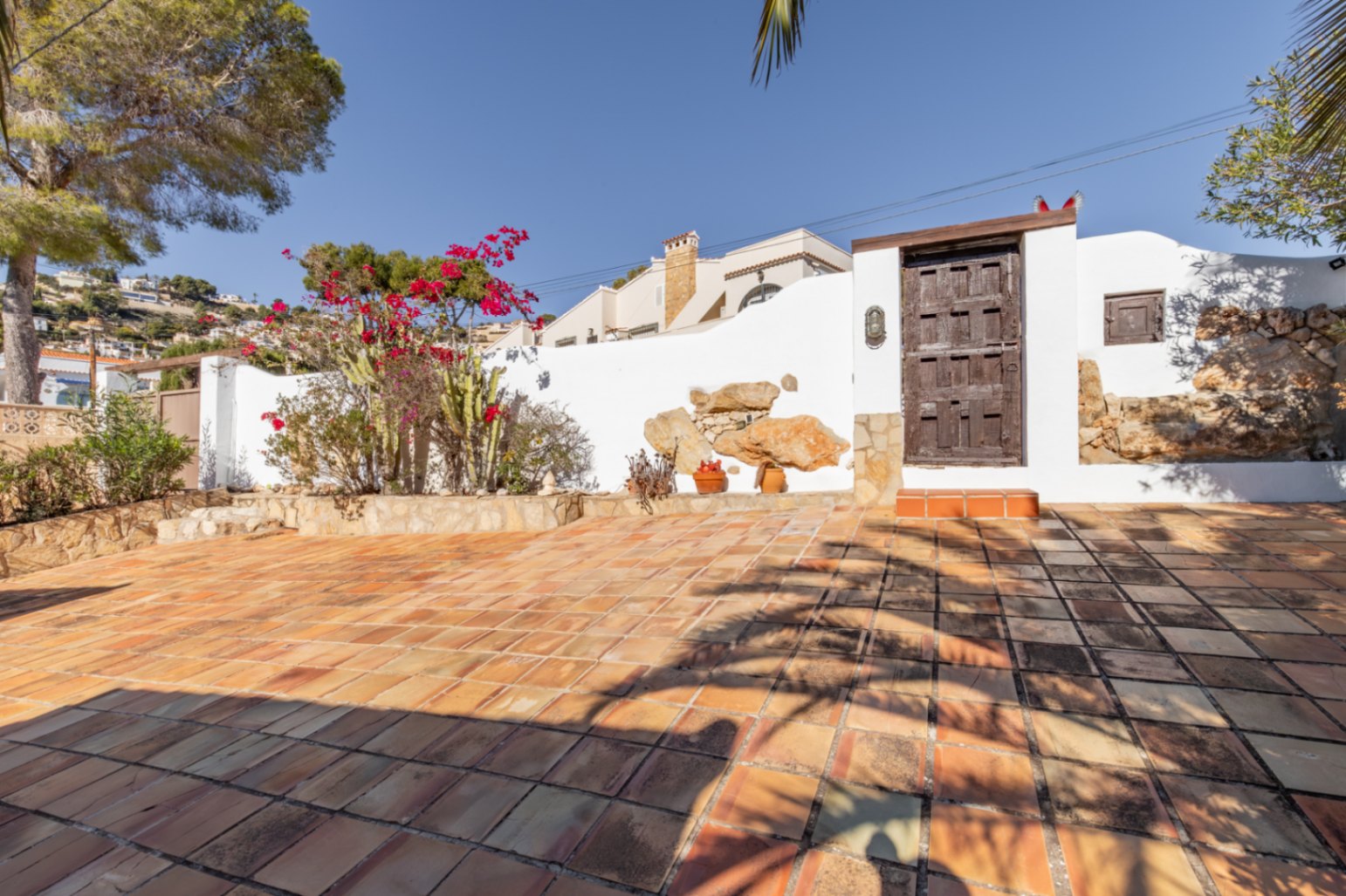 Outdoor terrace featuring terracotta tiling, white perimeter walls with natural stone inserts, and a rustic wooden gate framed by Mediterranean bougainvillea and cacti.