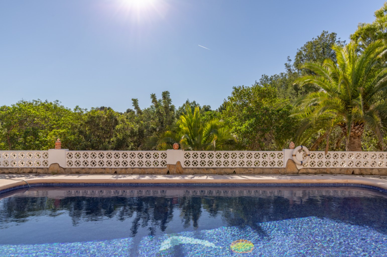 South-facing swimming pool with blue mosaic tiling, bordered by a white geometric balustrade and mature Mediterranean trees under direct sunlight.