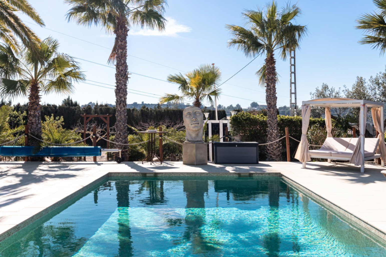 A 9.5m x 5m swimming pool featuring a manual cover, stone head sculpture, and a white Balinese daybed, all set within a landscaped terrace with mature palm trees.