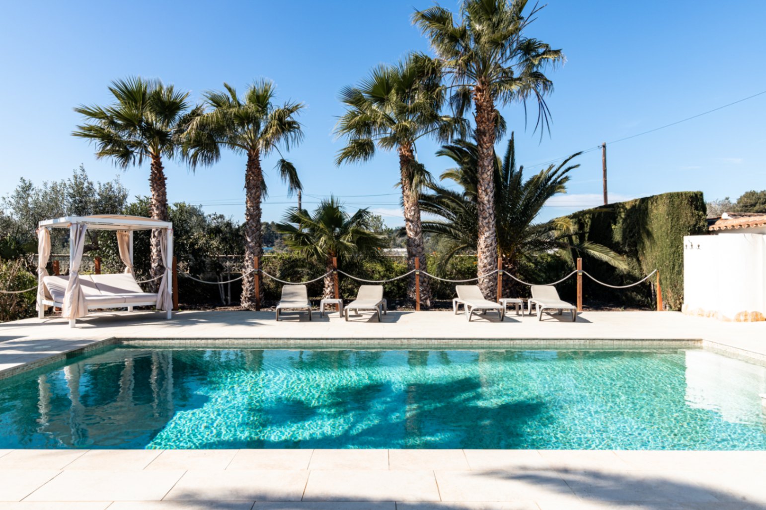 A 9.5m x 5m swimming pool featuring a paved sun terrace with four loungers, a curtained daybed, and a backdrop of mature palm trees.