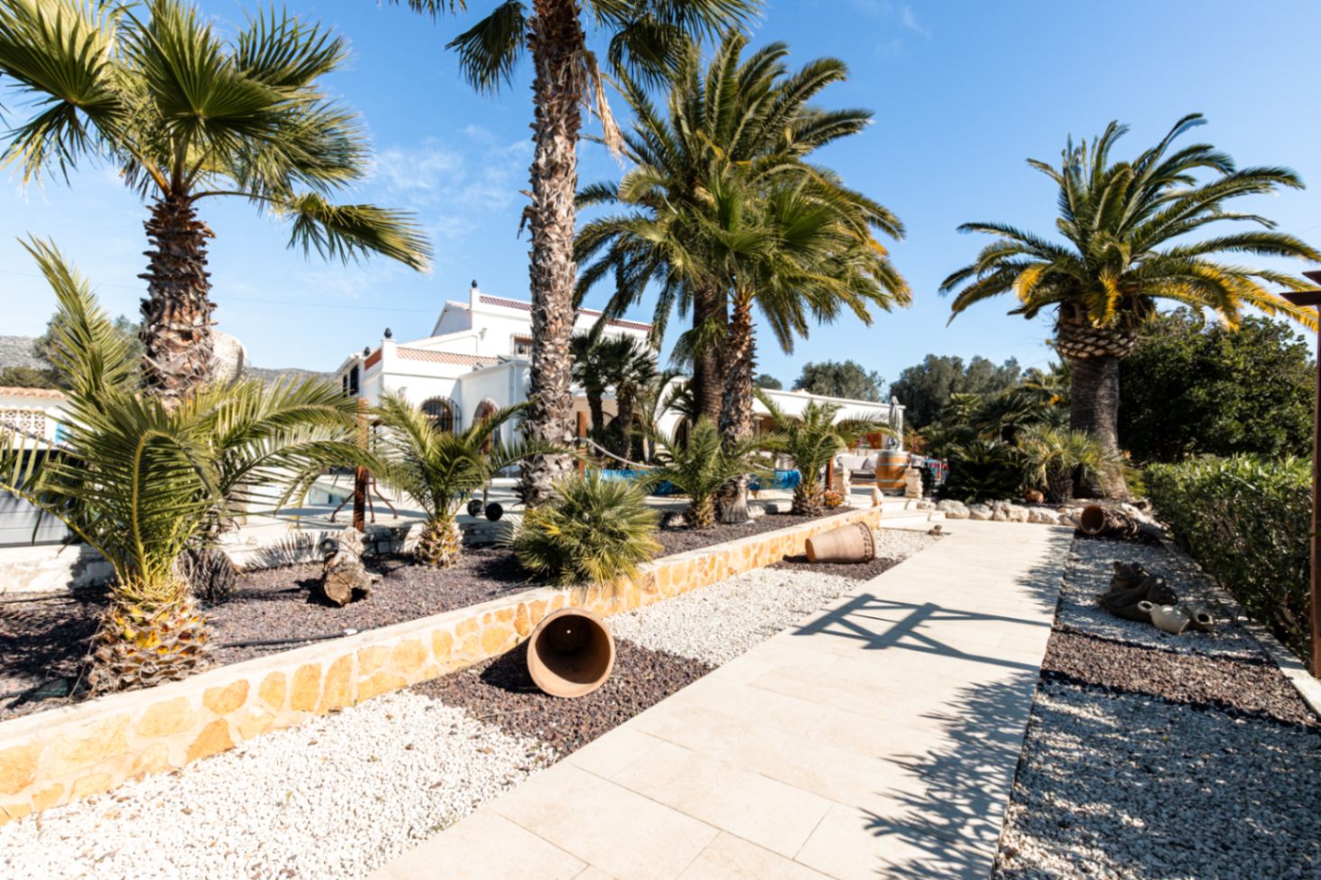 A light-tiled pathway winds through landscaped gravel gardens with mature palm trees and decorative terracotta pots, leading toward the white Mediterranean villa.
