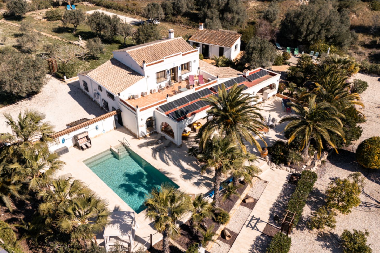 Aerial perspective of the finca showcasing the rectangular pool, stone terraces, and solar panel array atop the traditional naya, set within lush gardens.